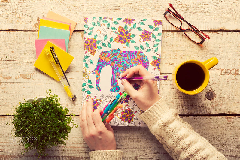 Overhead view of a finished fantasy coloring page showing a beautifully colored dragon with rich jewel tones and metallic effects, colored pencils arranged by color family nearby, satisfying completed artwork, warm dramatic lighting