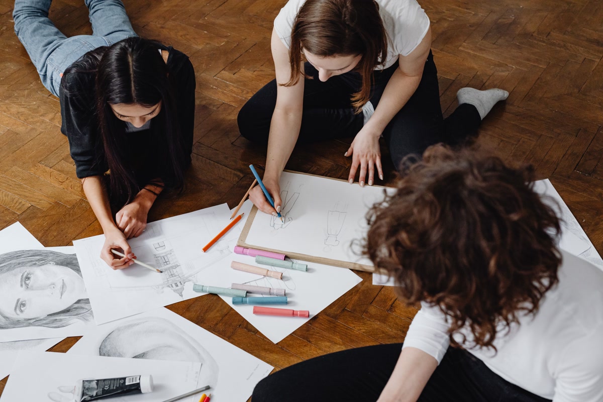 Close-up of multiple coloring books open side by side on a table with different people coloring, hands with different colored pencils visible, warm social atmosphere, diverse group of adults enjoying a shared creative activity