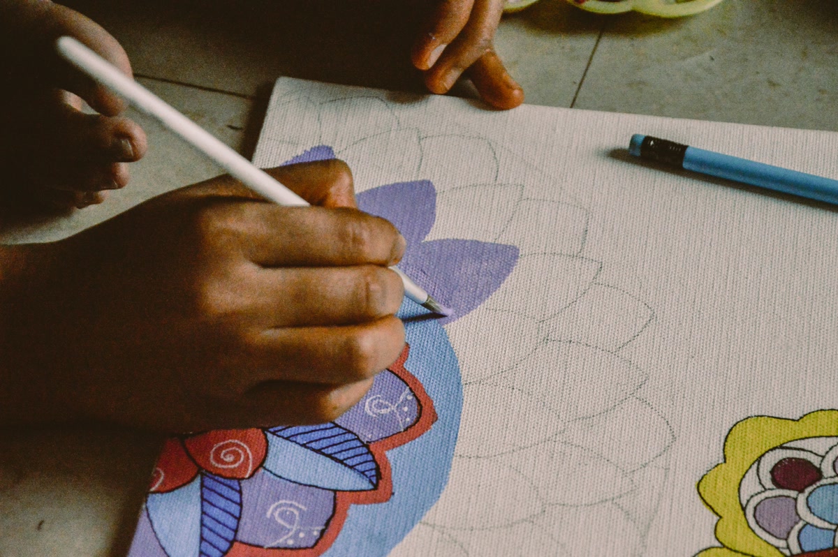 Close-up of hands coloring a mandala design with colored pencils, working from the center outward, vibrant colors laid down in symmetrical sections, warm focused lighting, meditative and precise technique, shallow depth of field