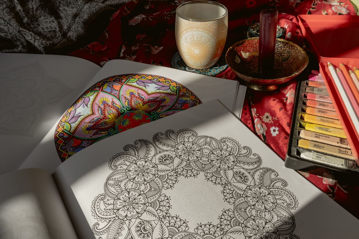 Hands coloring a vibrant floral mandala with warm orange and yellow colored pencils, bright colors contrasting with a gray winter window behind, close-up lifestyle photography showing the joy of color during winter